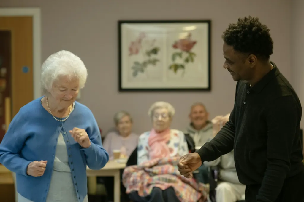 An older woman laughs and dances with a caregiver in a bright common room