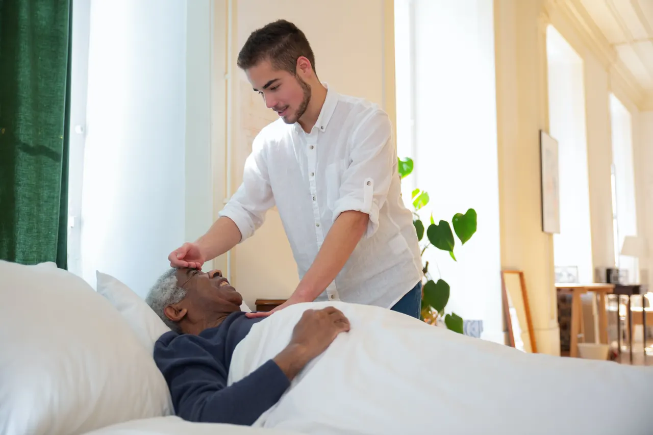 A caregiver checks on an older man resting comfortably in bed at home