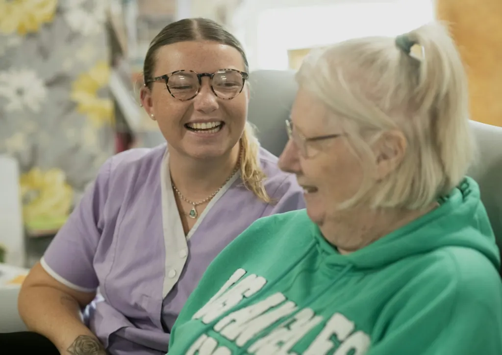 A smiling caregiver shares a laugh with an older woman in a green sweater