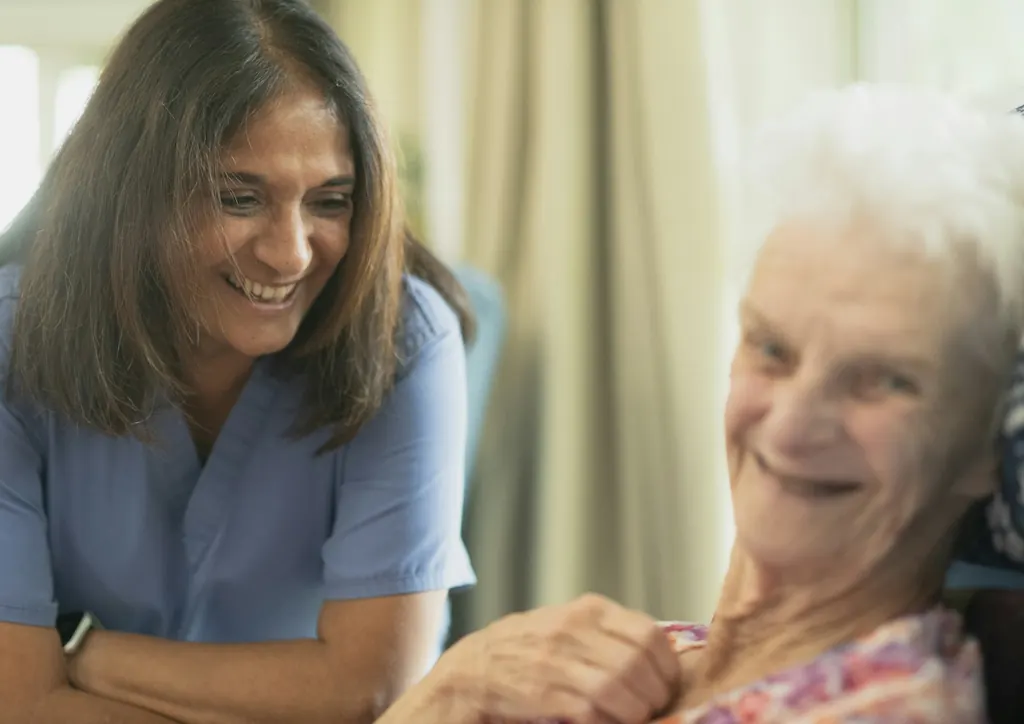 A nurse in blue scrubs smiles with an older woman during a visit