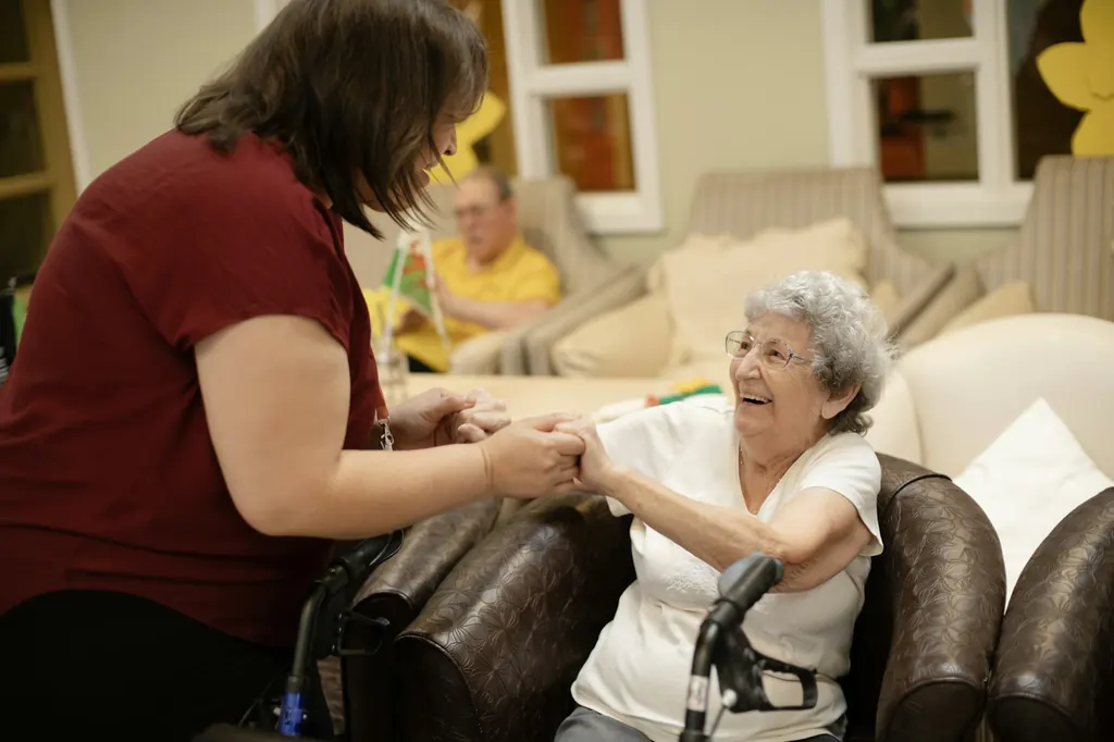 A caregiver helps an older woman stand with her walker in a sunny living room