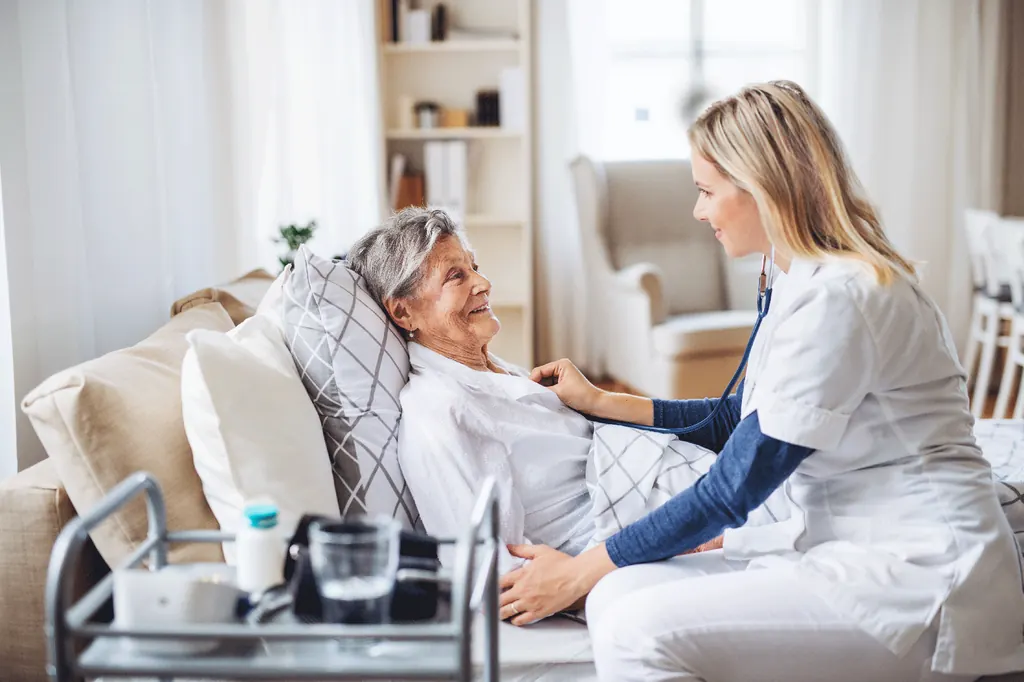 A nurse sits beside an older woman resting in bed at home, both smiling