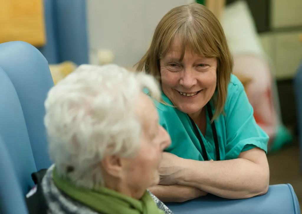 A smiling therapist in green scrubs leans in to talk with an older woman
